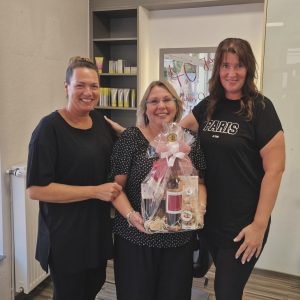 3 Frauen lachen in die Kamera. Die Frau in der Mitte hält ein Präsent in den Händen für ihr Betriebsjubiläum.