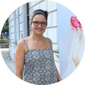 Romy Schmidt steht draußen in einen schönen Kleid mit Muster in Grau. Die Arme sind frei weil es warm ist. Sie steht und lacht in die Kamera