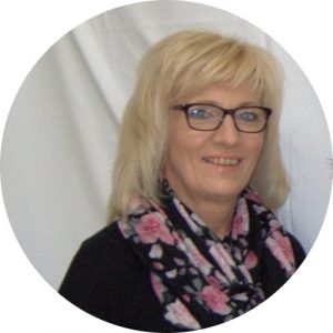 Eine Blonde Frau mit Schulterlangen Haare und schwarzen pullover mit schal. Sie steht vor einen weißen hintergrund und lächelt