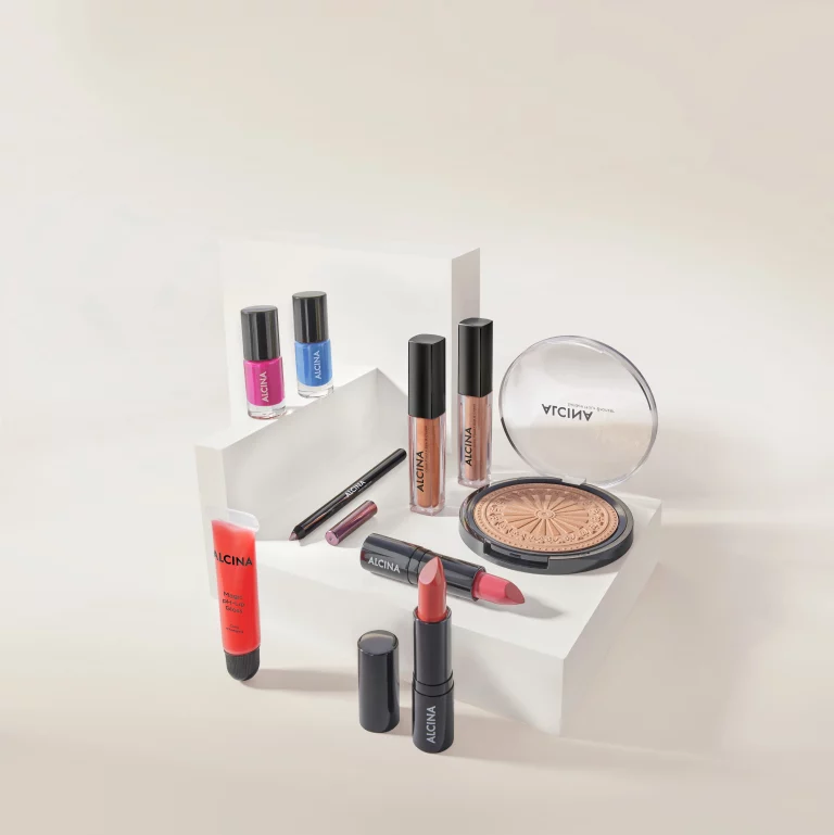 Ausgewählte ALCINA Make-up Produkte dekorativ auf weißen Podesten arrangiert. Zu sehen sind Nagellacke in Pink und Blau, Lipglosse, Lippenstifte, ein Lipliner, der Magic pH-Lip Gloss sowie der Golden Hour Bronzer mit geöffnetem Deckel in einem hellen, minimalistischen Setting