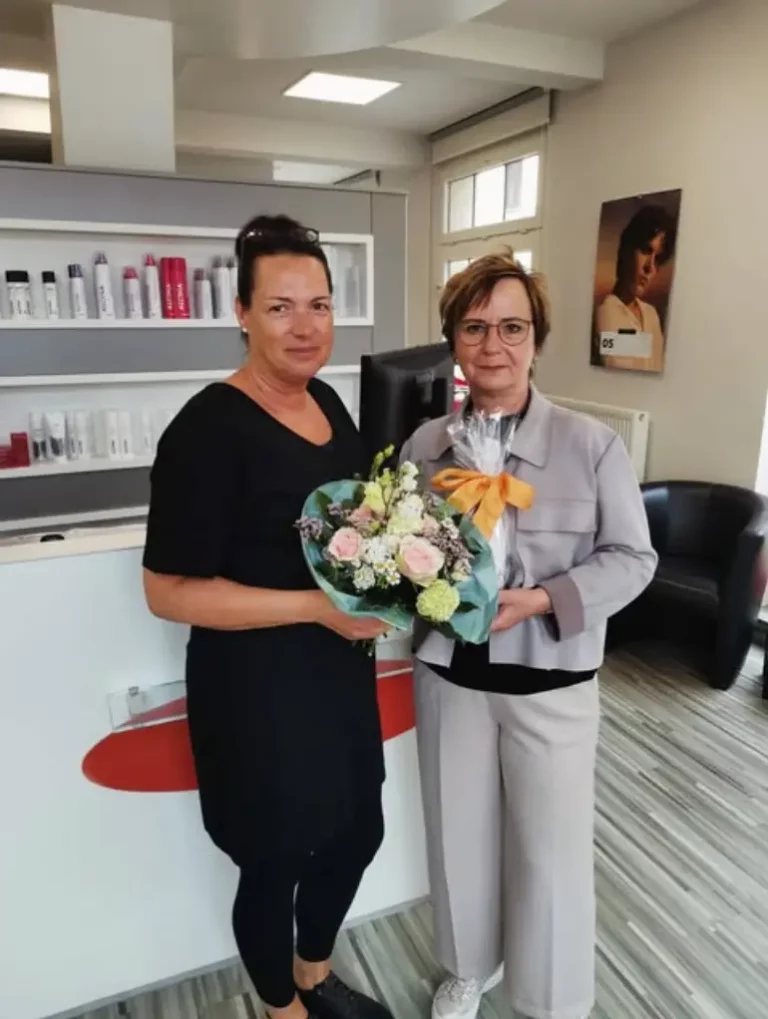 zwei Friseur Kolleginnen stehen im Salon in der Steinstraße. Überreicht wird ein Geschenk mit Blumenstrauß.